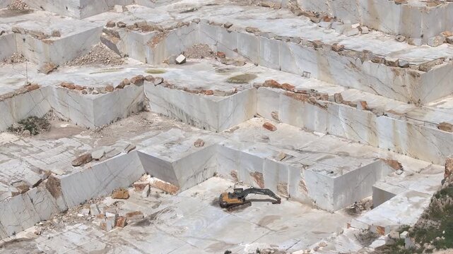 SICILY, ITALY, 24 JAN 2026: AERIAL, CLOSE UP: Excavator sits on a bright marble bench surrounded by huge cut walls and scattered stone blocks. Modern quarry operations and marble industry in Sicily.