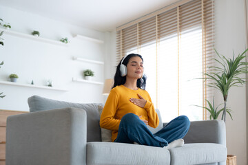 Obraz premium Happy young Asian woman practicing yoga and meditation at home sitting on couch in living room in lotus position and relaxing with closed eyes. Mindful meditation and wellbeing concept