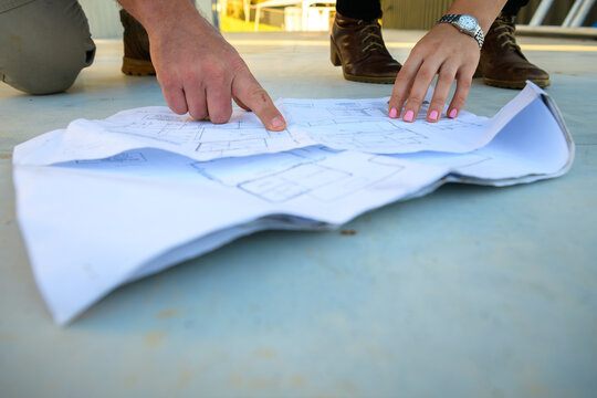 A close-up, low-angle shot of two people reviewing architectural blueprints laid out on a concrete floor. Ideal for construction planning, architectural design, and project management themes.
