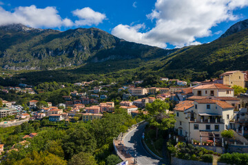 Fototapeta premium Picturesque view of Maratea with sunlit houses and terracotta rooftops nestled against lush green mountains. Dramatic shadows, vibrant foliage, and a clear blue sky create a tranquil Mediterranean