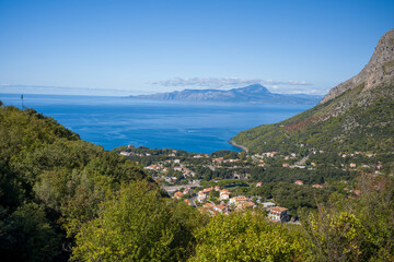 Fototapeta premium Elevated view of a coastal Italian village surrounded by dense green hills, overlooking the calm blue Tyrrhenian Sea and distant mountains under a clear sky. Bright sunlight enhances the vibrant