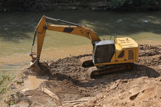 Powerful yellow excavator digging mud from riverbank. Heavy machinery at construction site, industrial equipment busy with earthmoving work on waterway
