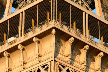 Close-up of the Eiffel Tower's ornate platform corner, bathed in warm golden sunlight, highlighting detailed ironwork, decorative corbels, and textured surfaces. © Florent