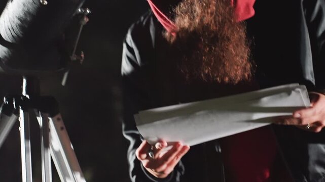 night backyard astronomer unfolding star charts, preparing telescope for stargazing session candid lowlight shots of hooded observer handling papers, checking notes and adjusting gear under soft