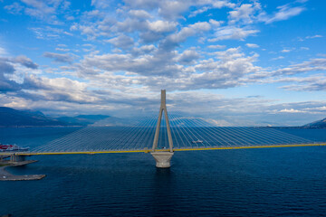 Obraz premium Modern cable-stayed Rion-Antirion Bridge spans deep blue water near Patras, Greece, with dramatic clouds and distant mountains under a vivid sky. The geometric lines of the bridge create a striking