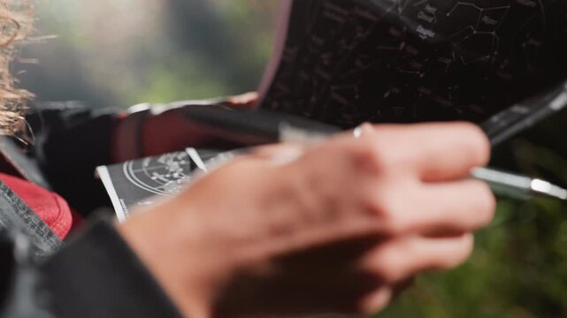 hands holding star chart marking constellations outdoors, closeup of hands annotating celestial map with pen, sunlight through foliage creating soft bokeh, researcher preparing telescope alignment