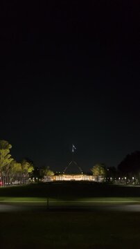 Vertical shot of Australian Parliament House facade illuminated at night with Australian flag flying in breeze atop flagpole in Canberra. National landmark lit up vertical format for social media