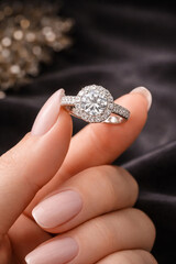 Close-up luxury diamond ring held between fingers, featuring a brilliant round-cut diamond with a halo setting and pav&eacute; diamond band. Shot on a dark elegant background,