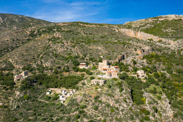 Fototapeta premium Aerial view of traditional stone buildings nestled among rocky slopes and green vegetation on a hillside in Mani, Greece, under a clear blue sky.