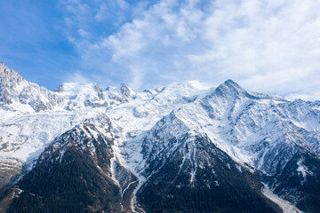 Obraz premium Expansive view of the Mont Blanc massif with rugged snow-covered summits and forested lower slopes, illuminated by soft daylight beneath a partly cloudy sky.