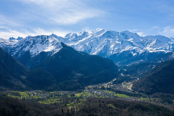 Fototapeta premium Aerial view of Les Houches village nestled in a green valley, with the dramatic snow-capped peaks of the Mont Blanc massif rising in the background under a clear blue sky.