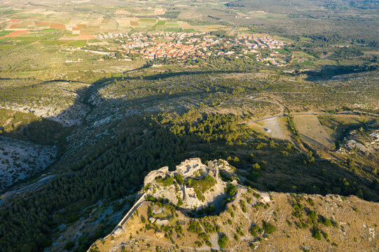 Ancient stone ruins of Chateau de Opoul Perillos crown a rugged hilltop, overlooking a patchwork of fields and the distant village under warm sunlight in southern France.
