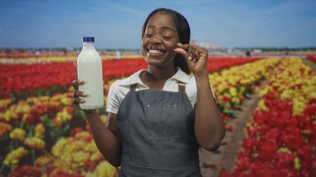 Woman in denim apron holding milk bottle with blue cap, smiling and pinching fingers near thumb in studio among tulip field backdrop; simple joy.