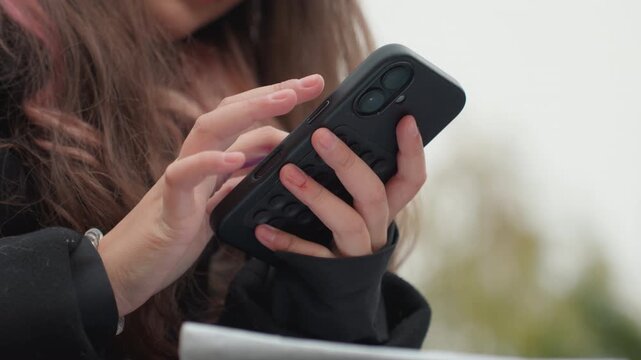 young caucasian student using smartphone outdoors, closeup of hands typing and scrolling on black case, long hair and winter coat visible, checking messages and composing notes for class, candid study