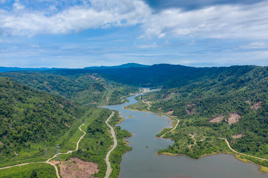 Aerial perspective of a river meandering between lush, densely wooded hills and patches of cleared land under a bright sky. The landscape features winding water, vivid greenery, and natural curves in