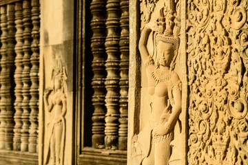 Warm sunlight illuminates detailed sandstone carvings of apsara dancers and ornate floral motifs on the temple wall at Angkor Vat, Cambodia. The intricate Khmer reliefs are framed by textured stone © Florent
