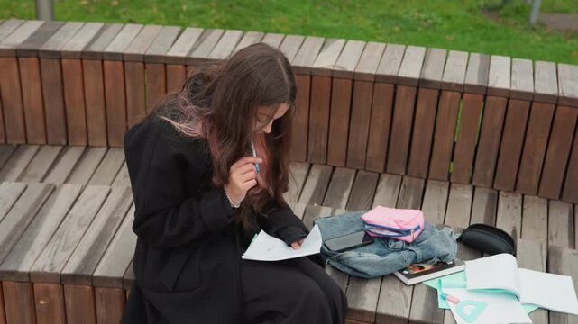 student studying on bench with scattered papers, chewing pen while reading notes, backpack and supplies beside seat, intense revision for exam, overcast park setting, candid campus study moment,