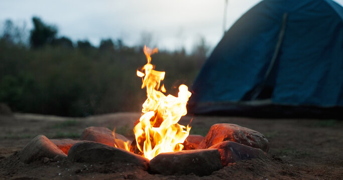 Night, tent and wood with flames for bonfire, smoke and embers burning for camping travel holiday. Evening light, heat source and glowing log with sparks in nature, rocks and outdoor for weekend trip