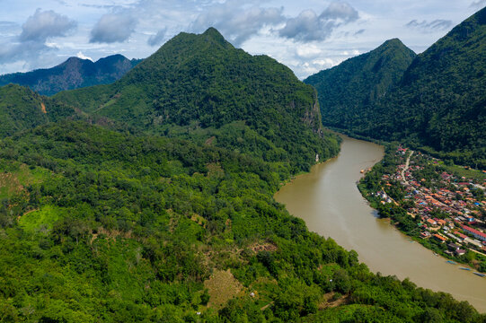 Aerial view of the Nam Ou river curving between lush forested mountains and the village of Nong Khiaw in northern Laos. Dense tropical vegetation and dramatic limestone peaks create a vibrant
