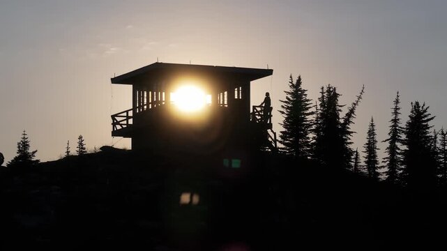 A panning aerial drone shot moving from right to left showing a silhouetted remote isolated fire tower lookout in remote northern Idaho as the sun passes through windows and a woman leans on a railing