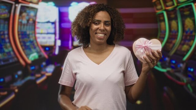Woman holding heart box and showing four fingers in casino building wearing pink tshirt and smiling broadly; happy celebration.