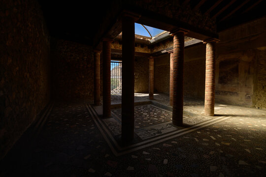 A shadowed Roman atrium with brick columns and a central impluvium is illuminated by natural light from an open skylight in the House of Menander, Pompeii, Italy. Mosaic flooring and textured stone