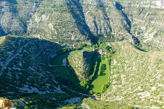 Sweeping aerial view of a lush green valley winding through steep limestone hills at Cirque de Navacelles. Bright sunlight enhances the vivid contrast between fertile meadows and rugged rocky terrain.