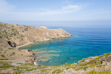 A tranquil, rocky inlet with transparent turquoise water and sparse Mediterranean vegetation along the rugged coastline of Asinara National Park, Sardinia, under a soft blue sky.