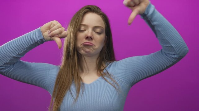 Young caucasian woman with straight long hair and bare thumb pointing down in purple studio wearing blue top; rejection.
