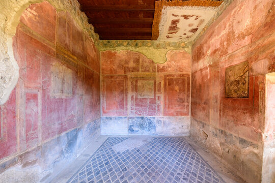 Ancient Roman room in the House of Menander, Pompeii, Italy, featuring faded red frescoed walls and a geometric mosaic floor. Sunlight highlights the weathered plaster, textured surfaces, and remnants