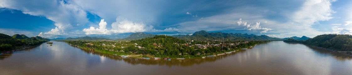 Fototapeta premium Sweeping panoramic view of the Mekong River curving around the lush green hills and town of Luang Prabang, Laos. Dramatic clouds fill the expansive sky above forested mountains and riverside buildings