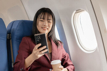Fototapeta premium A young woman sits by the airplane window holding a smartphone and a cup of coffee, enjoying a quiet moment, modern travel comfort, and relaxed personal time during a smooth flight.