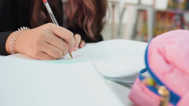 Focused student unspecified race writing notes closeup, hands holding pencil over open notebook, pink pouch in foreground, bracelet detail, soft bookshelf background, calm studious atmosphere,