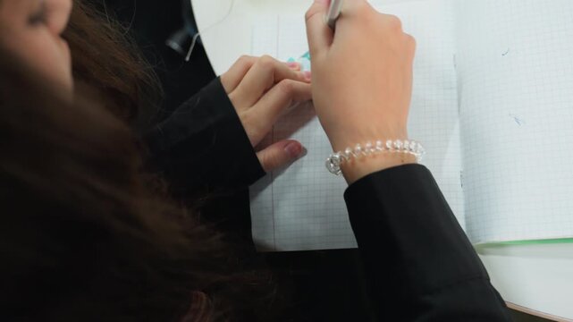 Student using protractor and pencil, female student drafting angles on grid paper at desk, closeup of hands with bracelet, focused study session with yellow cup in background, overhead classroom shot