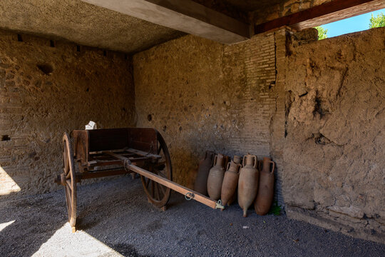 A rustic Roman storeroom in the House of Menander, Pompeii, Italy, features a weathered wooden cart and a group of terracotta amphorae against textured stone walls. Soft daylight highlights the