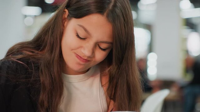 student girl focused at table studying, modern communal workspace with blurred background people and soft lights, long brown hair, pen and eraser in hand, flipping notebook pages, subtle smile