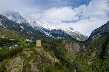 Obraz premium A solitary medieval stone tower stands atop a rugged ridge surrounded by dense forest, overlooking dramatic snow-capped peaks in Val d'Aoste, Italy. The landscape features textured rock, alpine