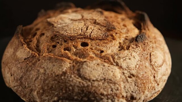Close-up detail of a freshly baked rustic sourdough bread loaf with a crispy, golden-brown crust against a dark background, emphasizing texture and detail.