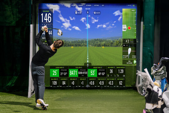 Indoor golf practice area featuring a golfer swinging a club towards a digital screen displaying performance metrics and a virtual landscape