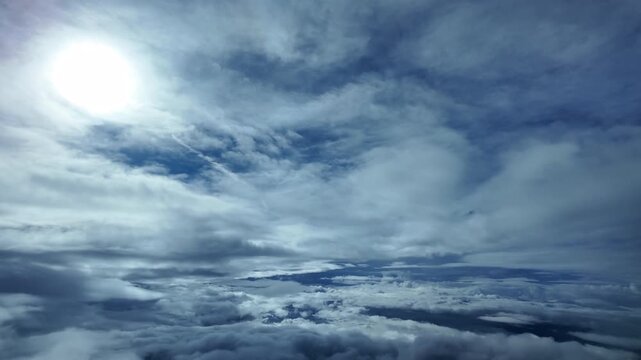 Immersive pilot view captured from the cockpit of a jet airplane flying at supersonic speed through layers of ethereal stratus clouds in a blue sky, with a veiled faded sun shaning above