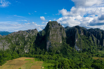 Obraz premium Towering limestone cliffs rise above green forest and patchwork fields under a vivid blue sky near Vang Vieng, Laos. Bright sunlight and scattered clouds highlight the dramatic vertical rock faces and