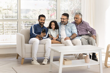 Fototapeta premium Small boy spend time with dad, grandfather and great-grandfather using smartphone sit together on sofa, look at device screen, make selfie pictures, engaged in videocall talk with family living abroad