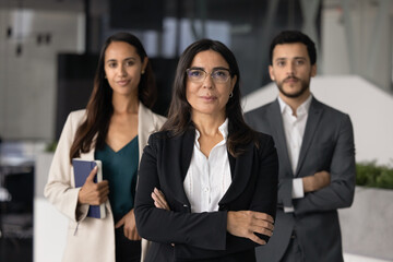 Mature 60s businesslady boss and two young 30s multiethnic subordinates standing in office, representing professional company services. Accounting or marketing department posing for photo at workplace