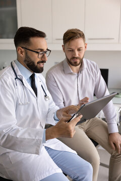 Trusted medicine. Confident young doctor in lab coat check paper healthcare records review personal medical chart in presence of patient answer questions explain test results discuss treatment options