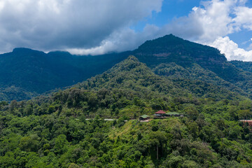 Naklejka premium A small lodge with red and green roofs sits nestled in dense tropical forest on a verdant hillside, with dramatic mountain peaks and low clouds in the background. The scene captures the rich greenery