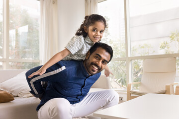 Obraz premium Happy Indian man enjoy funny active games with little son indoor, posing, looking at camera seated with child on sofa. Cute preschooler boy piggyback daddy, spend carefree playtime together at home