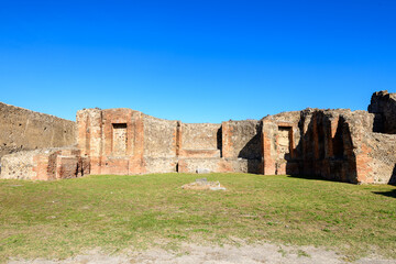 Ancient brick and stone remains of the Sanctuary of the Public Lares stand beneath a vivid blue sky in Pompeii, Italy. Sunlight highlights the textured masonry and open grassy courtyard, evoking the