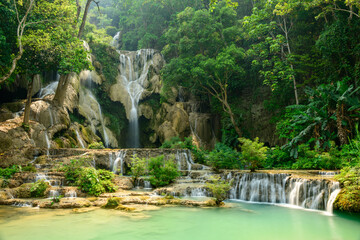 Lush green forest surrounds the multi-tiered limestone cascades and turquoise pools of Kuang Si Waterfalls in northern Laos. Sunlight highlights the flowing water and vibrant foliage, creating a © Florent