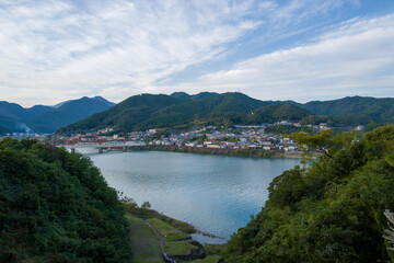 A tranquil riverside town sits at the base of lush green mountains, with a calm river curving through the landscape under a wide, softly clouded sky in Shingu, Japan.
