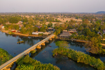 Fototapeta premium Aerial perspective highlights the historic French colonial bridge connecting Don Det and Don Khon in the 4000 Islands, southern Laos. Sunlit village houses, lush greenery, and tranquil river water